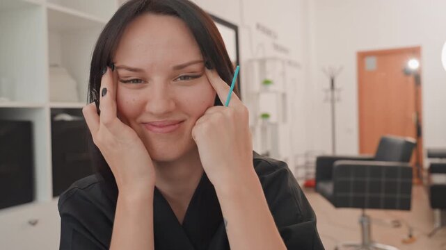 female stylist demonstrating eyebrow brush technique with playful smile and direct eye contact in modern salon, closeup on face and tool, ringlight glow and studio chairs in background beauty