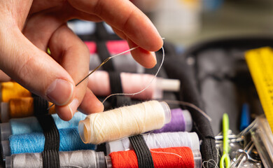 Gros plan sur une main en train de mettre du fil dans une aiguille. En arrière plan, une trousse à couture avec des bobines de fil. © Gautier Normand