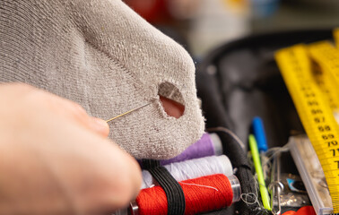 La main d'une personne en train de réparer un trou dans une chaussette avec une aiguille et du fil. En arrière plan, une trousse à couture avec des bobines de fil. © Gautier Normand