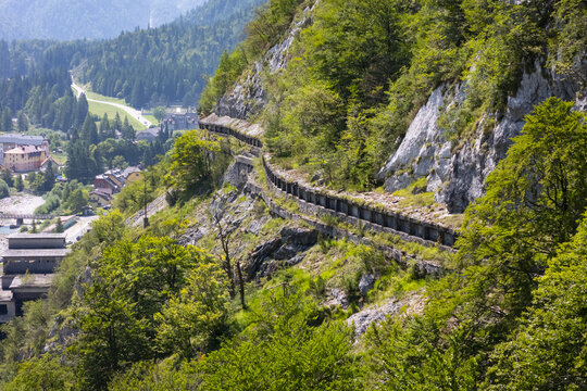 Aerial view of a winding path carved into a verdant mountainside, contrasting against the distant town nestled in the valley, Rabelj, Friuli-Venezia Giulia, Italy.