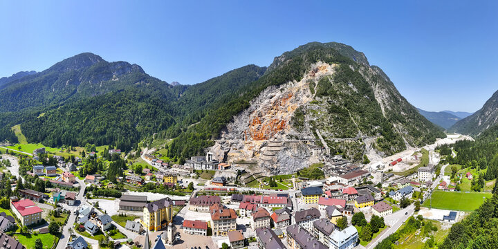 Aerial view of quaint village nestled in a valley, with a stark, terraced quarry scarring a verdant mountain, contrasting nature's beauty with industry's impact, Rabelj, Friuli-Venezia Giulia, Italy.