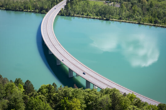 Aerial view of a graceful bridge arching over the turquoise Drava River, reflecting the sky amidst lush forests, Ferlach, Carinthia, Austria.