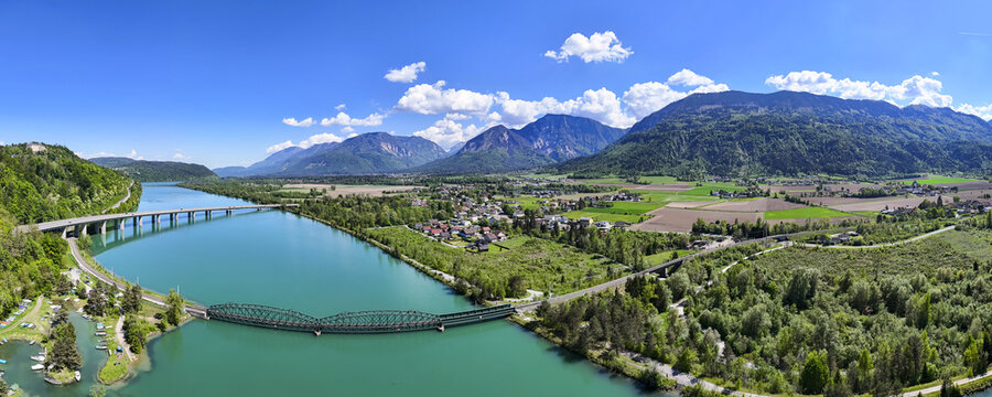 Aerial view of the turquoise Drava River reflecting the sky as it winds through green landscapes and under bridges, Ferlach, Carinthia, Austria.