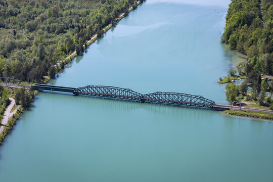 Aerial view of a symmetrical bridge spanning the turquoise Drava River, framed by lush green trees under the sunlight, Ferlach, Carinthia, Austria.
