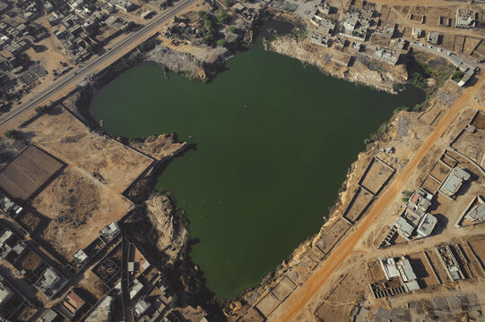 Aerial view of a dark green lake surrounded by arid land and scattered buildings under the bright sun, Gwazaye, Kano, Nigeria.