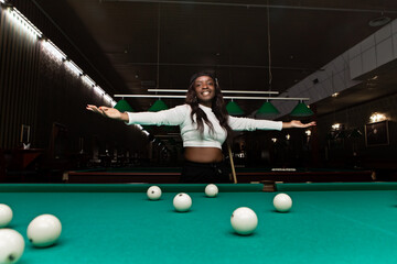 Young black woman wearing casual clothes, playing billiards, extending her arms, smiling and showing confidence in a pool hall, with green billiard table and white balls
