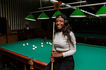 Smiling young black woman wearing a baseball cap posing with a cue stick on a green billiard table in a pool hall, enjoying a game of billiards