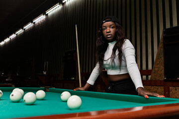 Young black woman with a baseball cap leaning on a pool table, starting a new game of russian billiards or pyramid, with white balls ready to be played