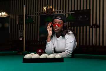 Trendy stylish woman wearing red sunglasses and baseball cap leaning confidently over pool table, concentrating during competitive game in elegant billiards lounge
