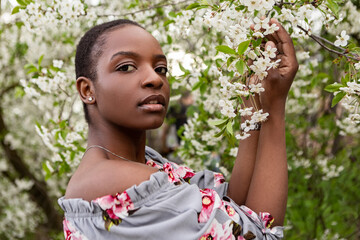 Young woman with short hair and floral off-the-shoulder top gently touching a branch of cherry blossoms in full bloom during springtime, enjoying the beauty of nature