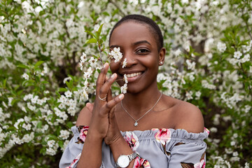 Young woman with short hair and floral off-the-shoulder top gently touching a branch of cherry blossoms in full bloom during springtime, enjoying the beauty of nature. Meditation, mental treatment
