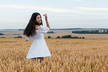 Young woman in a flowing white dress walking through a golden wheat field, smiling over her shoulder at the camera, with expansive farmland stretching out in the background