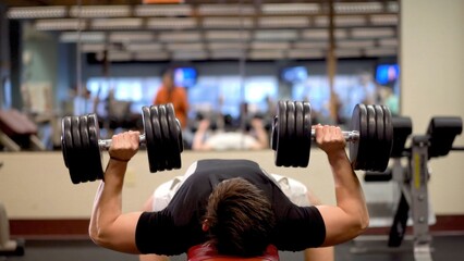 Young man performs strength training by lifting weights at gym while other people are working out in the background