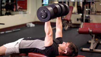 Obraz premium Young man performs weight training in gym using dumbbells while lying on bench to develop upper body strength and fitness