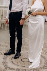 Bride in a white satin dress and groom in a classic shirt and tie standing side by side on a patterned carpet during a wedding ceremony