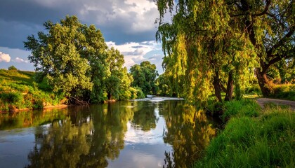 Serene river flanked by lush green trees, reflected light on water, grassy bank, and dramatic cloudy sky backdrop