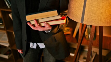 Person in dark suit holding a stack of old books near a floor lamp and bookshelf © shine.graphics