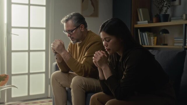 Grey-haired mature man and brunette girl both with their hands clasped in prayerful pose reading out prayer while sitting side by side on couch
