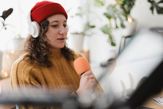 A young woman wearing headphones and a red beanie speaks into a microphone, recording audio in a home studio setting.