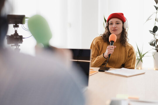 A woman wearing headphones and a red hat speaks into a microphone during a recording session, with a camera and laptop visible.