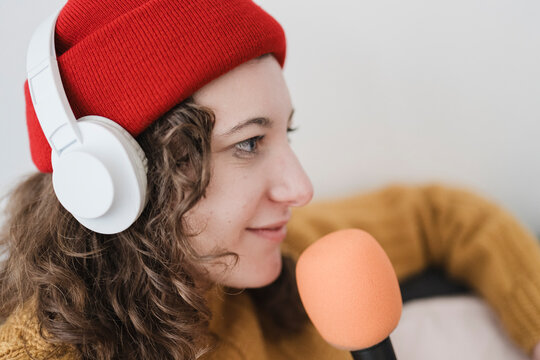 A young woman wearing headphones and a red beanie speaks into a microphone, creating content for a podcast or broadcast.