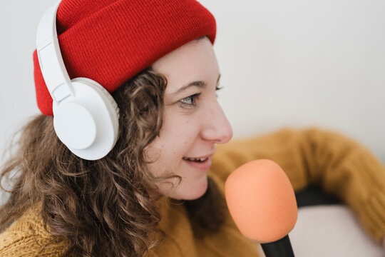 A young woman wearing headphones and a red beanie speaks into a microphone, recording audio for a podcast or broadcast.