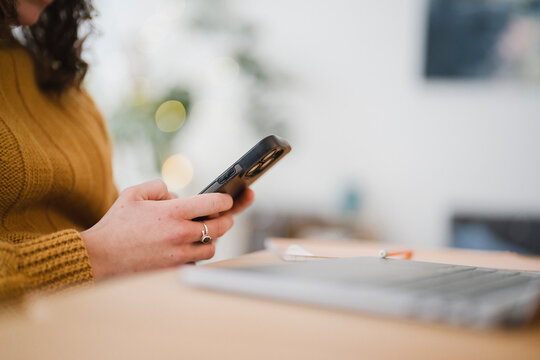 A person in a cozy sweater uses a smartphone while sitting at a desk with a laptop and papers.