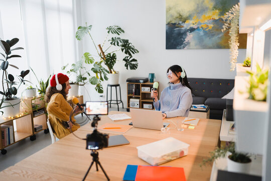 Two women with headphones are recording a podcast in a bright room, with microphones and a laptop on the table.