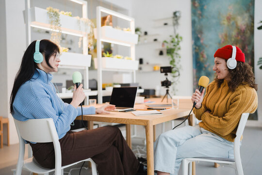 Two women wearing headphones sit at a table, each holding a microphone, engaged in a conversation or recording session.