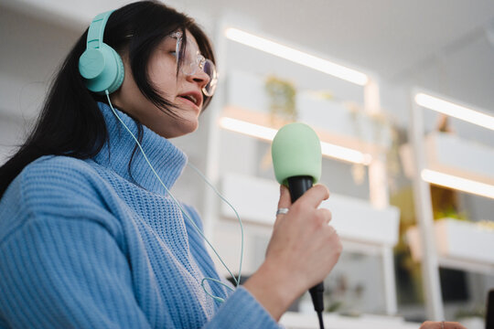 A young woman wearing headphones and glasses speaks into a microphone, recording audio for a podcast or broadcast.