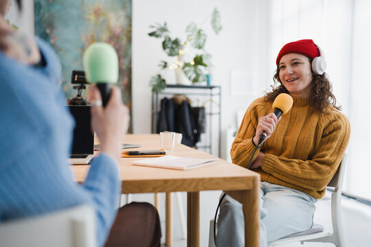Two people are recording a podcast interview in a bright studio, one holding a microphone and wearing headphones.