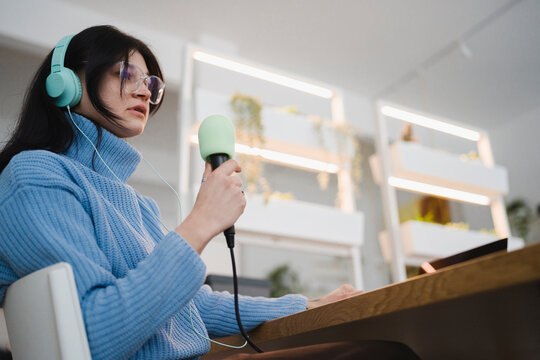 A young woman wearing headphones and glasses speaks into a microphone while sitting at a desk with a laptop.