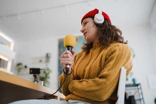 A young woman wearing headphones and a red beanie holds a microphone, recording audio in a studio setting.