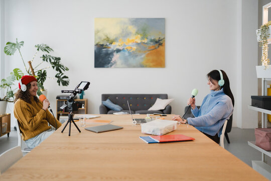 Two women wearing headphones are recording a podcast interview in a bright, modern room with plants and artwork.