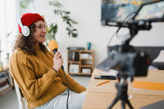 A young woman wearing headphones and a red hat speaks into a microphone while recording content with a camera in a home studio.