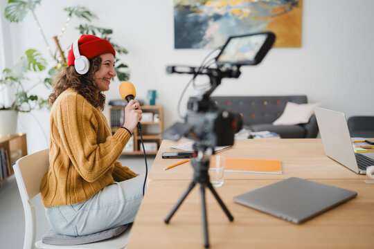 A young woman wearing headphones and a red hat records herself speaking into a microphone, with a camera and laptop set up for content creation.