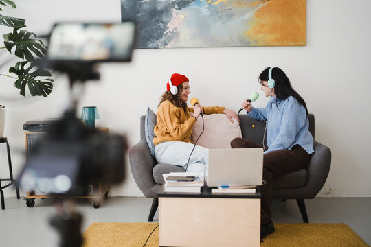 Two women wearing headphones are recording a podcast interview in a cozy studio setting, with microphones and a laptop visible.