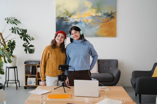 Two young women with headphones are setting up a camera and laptop for a video recording session in a bright, modern room.