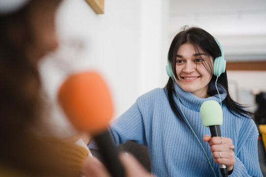 A young woman with headphones smiles while holding a microphone, engaging in a conversation or interview with another person.