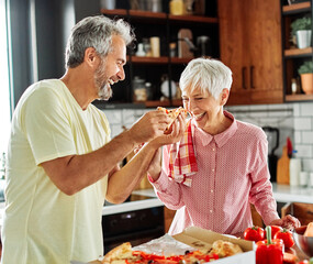 Portrait of happy senior couple prepering meal and tasting pizza in kitchen