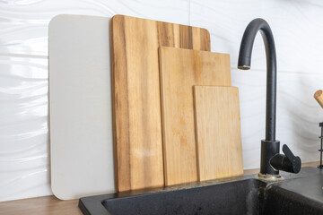 Kitchen cutting boards made of different materials standing near a sink. Wooden and plastic boards represent food preparation, hygiene, and safe cooking practices in a modern home kitchen.