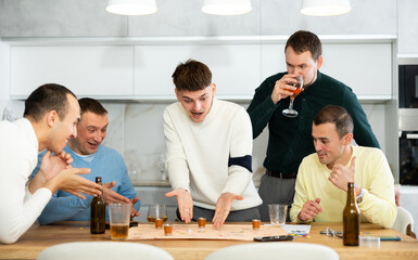 Carefree emotional male friends playing fun drinking board game with shot glasses replacing pieces during bachelor party at home..