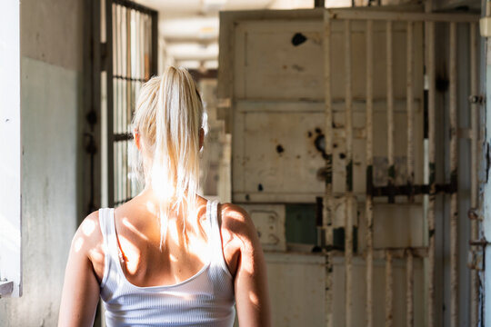 Blonde woman in white tank top seen from behind looking at old rusty metal bars and gates in abandoned prison corridor. - Powered by Adobe