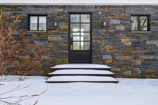 Modern stone house entrance with black front door and snow-covered steps, contemporary residential exterior in winter