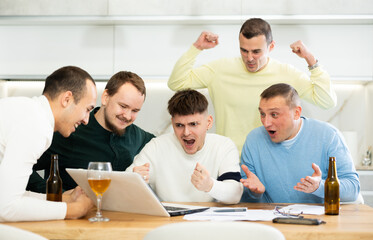Fototapeta premium Group of male friends gathering at home enthusiastically celebrating good news, seen on laptop screen, with lively gestures and drinks..