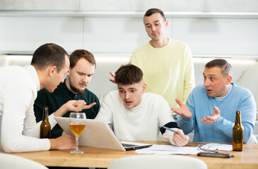 Obraz premium Group of men huddling around laptop at kitchen table, discussing problems and comforting distressed friend while gathering at home bachelor party
