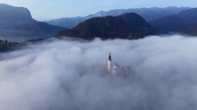 Aerial view of Lake Bled island church spire rising fom thick sea of clouds