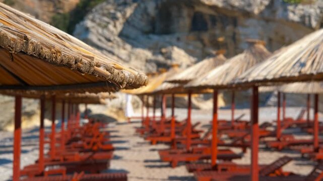 Empty beach with sunbeds and straw umbrellas. Empty red wooden sunbeds and straw thatched umbrellas standing in rows on a deserted sandy beach waiting for tourists on a sunny summer morning