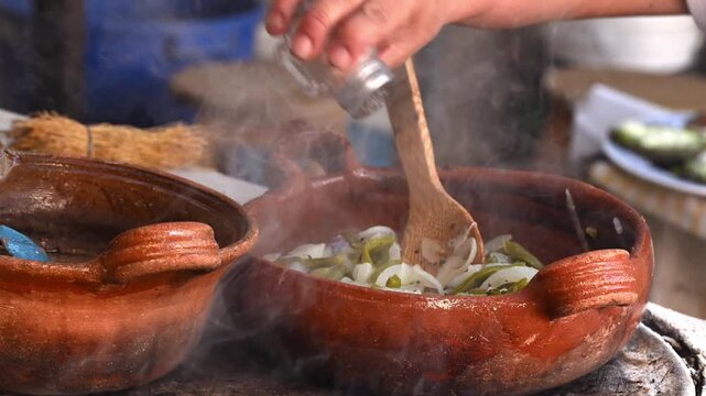 Close-up of hands adding salt to a clay pot with chopped cactus and onion