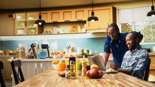 African American freelancer sitting at a kitchen table with a laptop, sending emails and explaining business tasks to his girlfriend. Couple enjoys a relaxed home lifestyle with coffee.